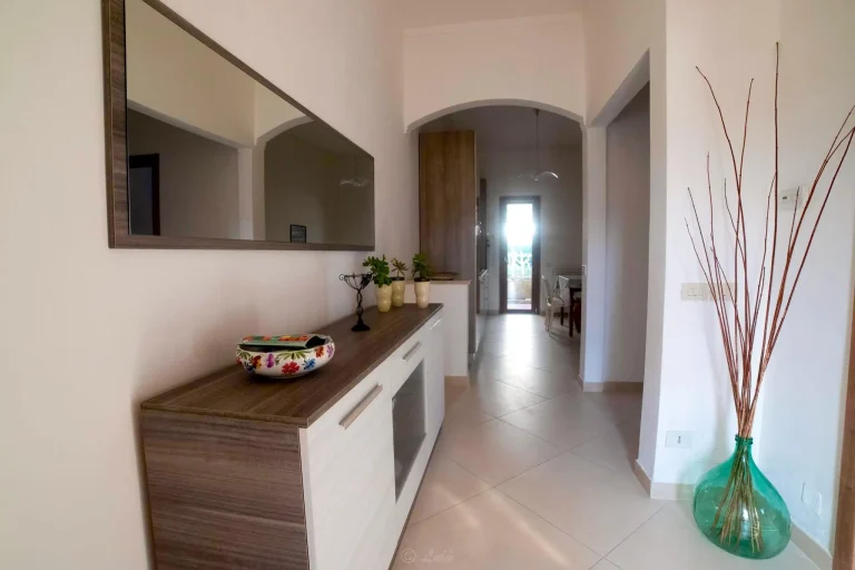 Pasillo moderno con piso claro, espejo grande en la pared, mueble con decoraciones, jarrón verde con ramas y puerta al fondo.