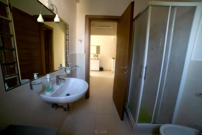 Baño con lavabo blanco, espejo grande, luz colgante, ducha con puertas de vidrio y puerta abierta hacia un pasillo iluminado.