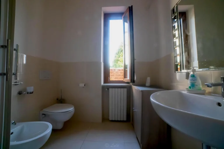 Baño con ventana abierta, lavabo moderno, espejo cuadrado, inodoro, bidé, y radiador bajo la ventana.