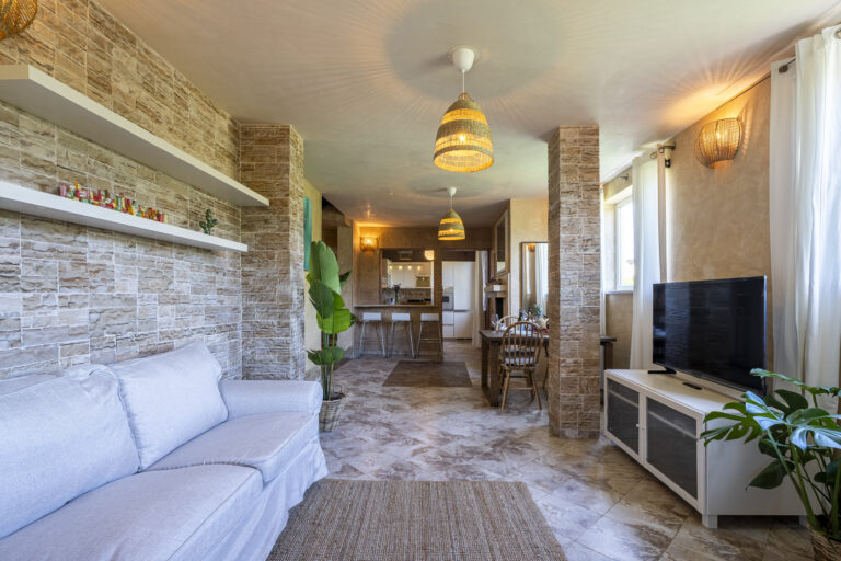 Sala de estar con sofá blanco, estantes flotantes, pared de piedra, plantas, televisor grande y lámparas colgantes.