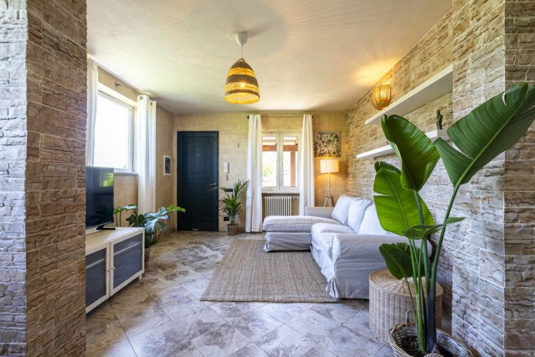 Sala de estar con paredes de piedra, sofá blanco en L, alfombra de yute, plantas y lámparas de mimbre iluminando.