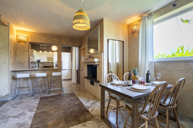 Cocina abierta con barra, taburetes blancos, mesa de comedor con sillas de madera, lámpara colgante, ventana con cortinas blancas.