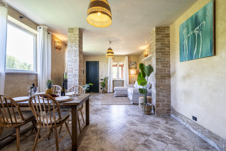 Sala moderna con comedor de madera, sillas de respaldo curvo, sofá blanco, lámparas colgantes, y plantas. Suelo de baldosas y paredes de piedra.