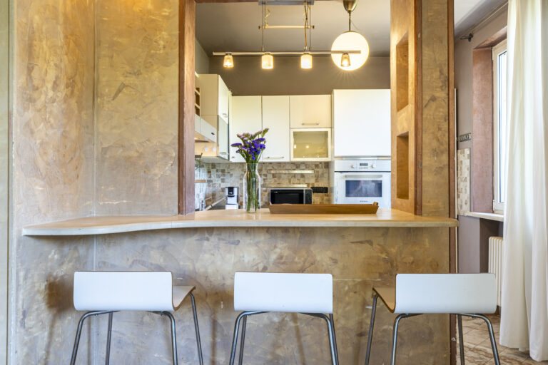 Cocina moderna con encimera, taburetes blancos, luces colgantes y paredes beige. Electrodomésticos blancos y flores en un jarrón.