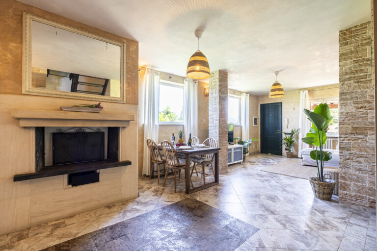 Sala luminosa con chimenea, mesa de comedor de madera, sillas, plantas, puerta azul y lámparas colgantes. Paredes y suelo de tonos neutros.