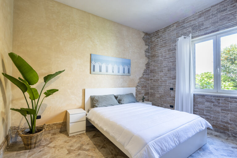 Habitación moderna con cama blanca, pared de ladrillos, planta en maceta, ventana grande y luz natural. Decoración minimalista.