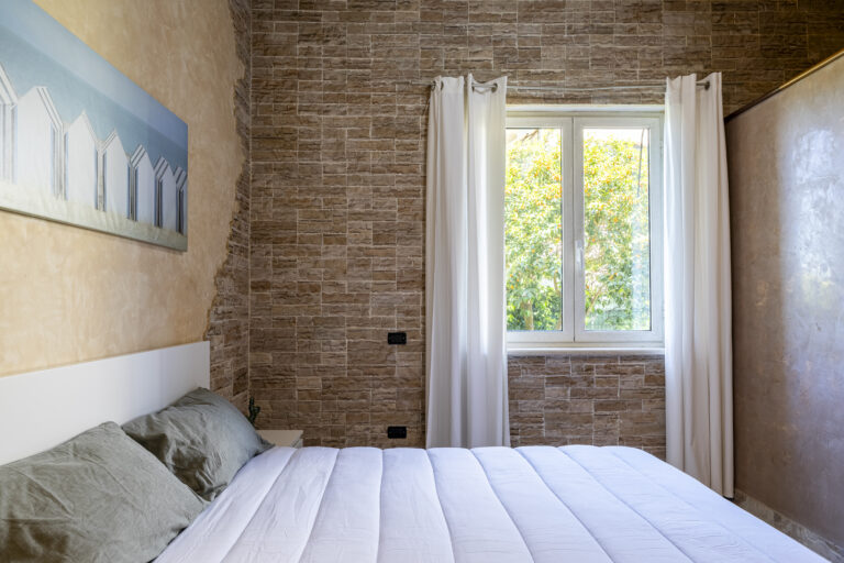 Dormitorio con pared de piedra y ventanas amplias, cama con edredón blanco, cortinas blancas y pared con pintura de edificios.