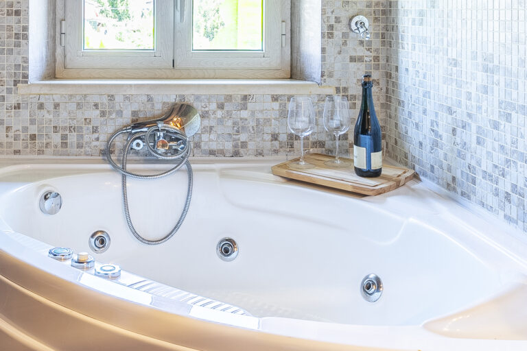 Bañera de hidromasaje blanca, azulejos de mosaico beige en la pared, botella de vino y copas sobre bandeja de madera junto a la ventana.