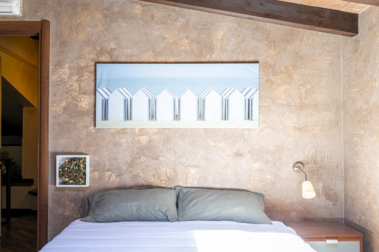 Dormitorio moderno con cama, almohadas gris oscuro, pared texturizada en tonos beige y cuadro de cabañas de playa. Iluminación suave.