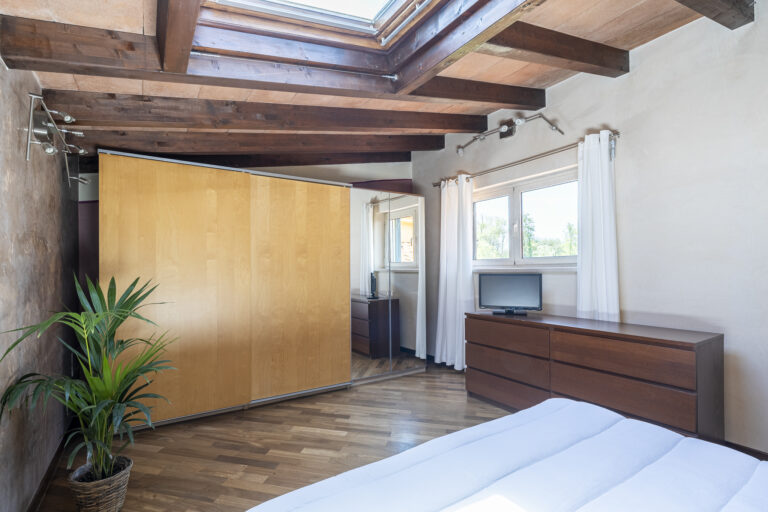 Habitación con techo de vigas de madera, cama blanca, armario grande, planta en maceta y ventanas que iluminan el espacio.