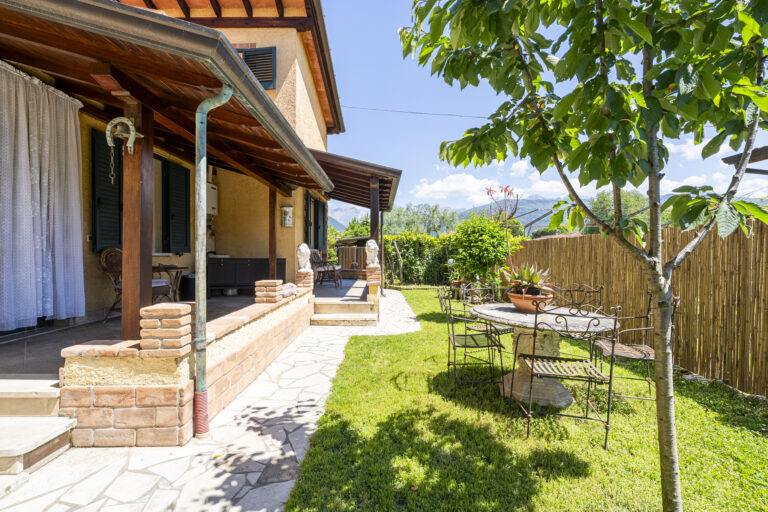 Casa de campo con porche de madera, jardín con césped, mesa de metal, sillas, árbol y fondo de montañas bajo cielo despejado.