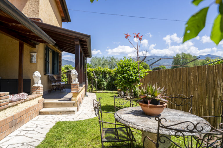 Casa rural con jardín verde, mesa de piedra, sillas de hierro, planta en maceta. Camino de piedra junto a la terraza de madera.