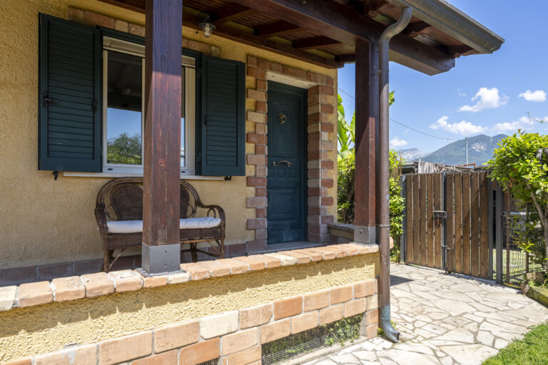 Pequeño porche de casa con puerta azul, paredes de ladrillo y ventana con contraventanas verdes. Silla de mimbre y camino de piedra.