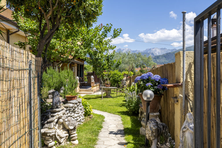Jardín con camino de piedra, cercado de bambú, plantas verdes, hortensias azules, estatuas y vista a montañas.
