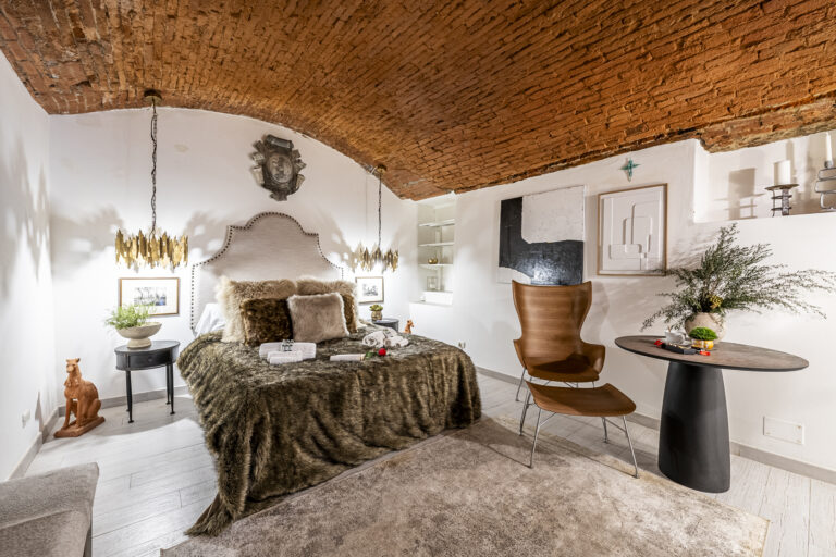 Habitación con cama de lujo, techo abovedado de ladrillo, lámparas doradas, arte moderno y silla elegante.