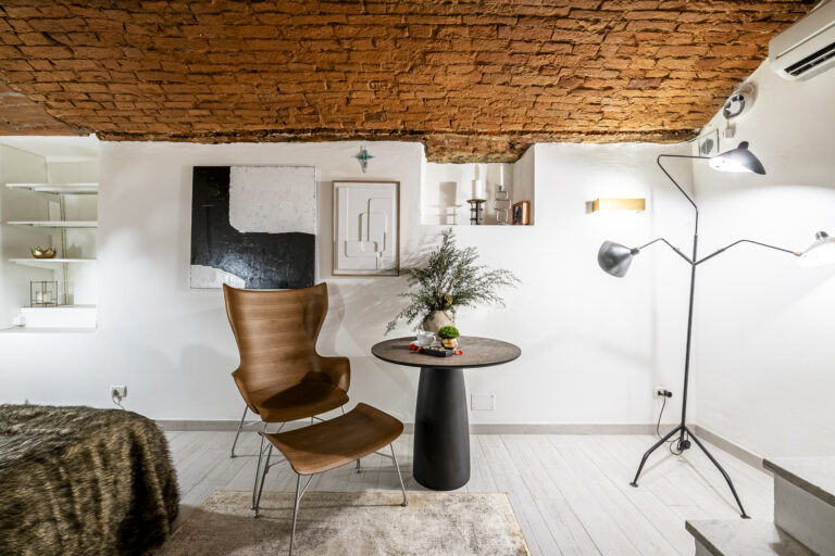 Habitación moderna con techo de ladrillo, silla de madera, mesa redonda, lámpara de pie y arte en pared blanca.