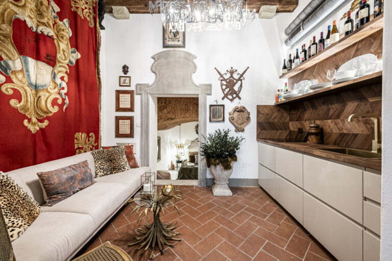 Sala con sofá beige, cojines de estampado animal, paredes decoradas con tapiz rojo y elementos artísticos. Cocina con estantes y vinos.