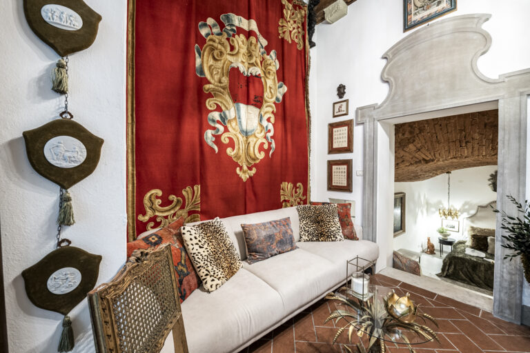 Sala con tapiz rojo y escudo dorado, sofá beige con cojines de leopardo. Mesa de centro dorada, puerta a habitación con techo abovedado.