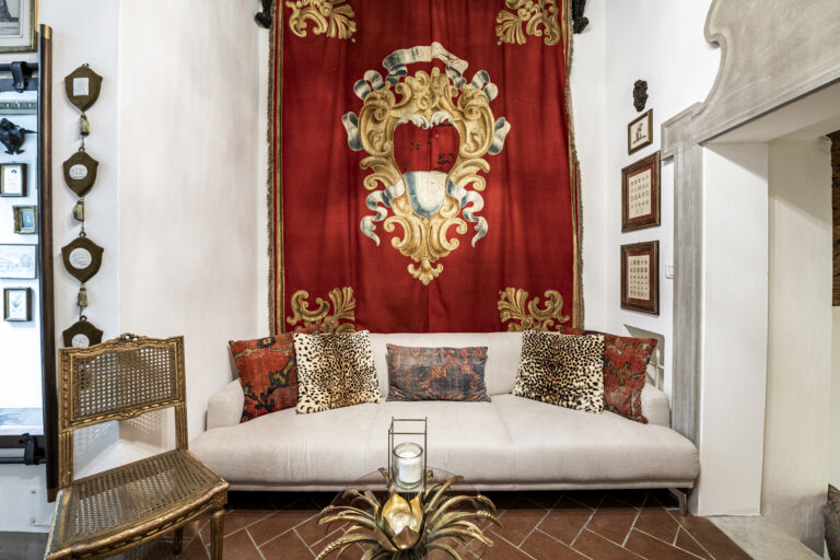 Sala de estar con sofá blanco, cojines estampados y tapiz rojo con escudo. Silla dorada y mesa con base decorativa.