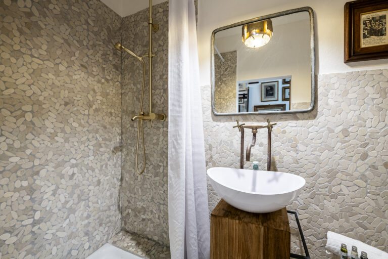Baño con paredes de mosaico de piedras, espejo rectangular, lavabo blanco sobre madera y ducha dorada.