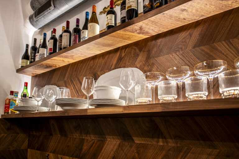 Estante de madera con copas, platos blancos, botellas de vino variadas y algunas botellas de licor. Ambiente cálido y acogedor.