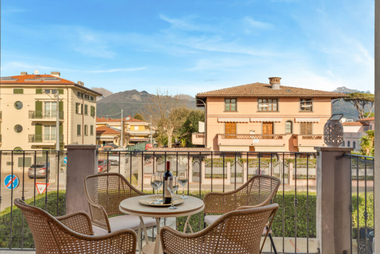 Terraza con mesas y sillas de mimbre, botella de vino y copas. Al fondo, edificios mediterráneos y montañas. Cielo azul claro.