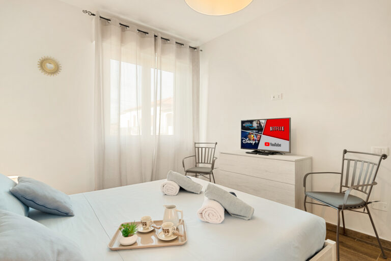 Dormitorio luminoso con cama de sábanas azul claro, TV con plataformas de streaming, dos sillas, ventana con cortinas blancas.