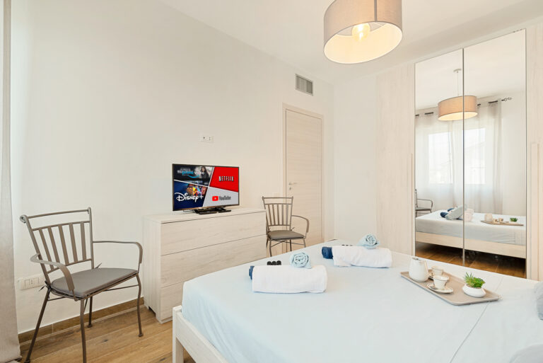 Habitación moderna con cama cubierta de sábanas azules, televisor en mueble blanco, armario con puertas espejo y decoración minimalista.