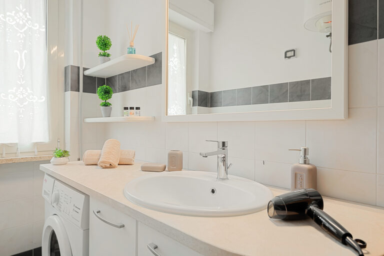 Baño moderno con encimera clara, lavabo blanco, grifo plateado, espejo grande, secador negro, toallas beige y plantas decorativas.