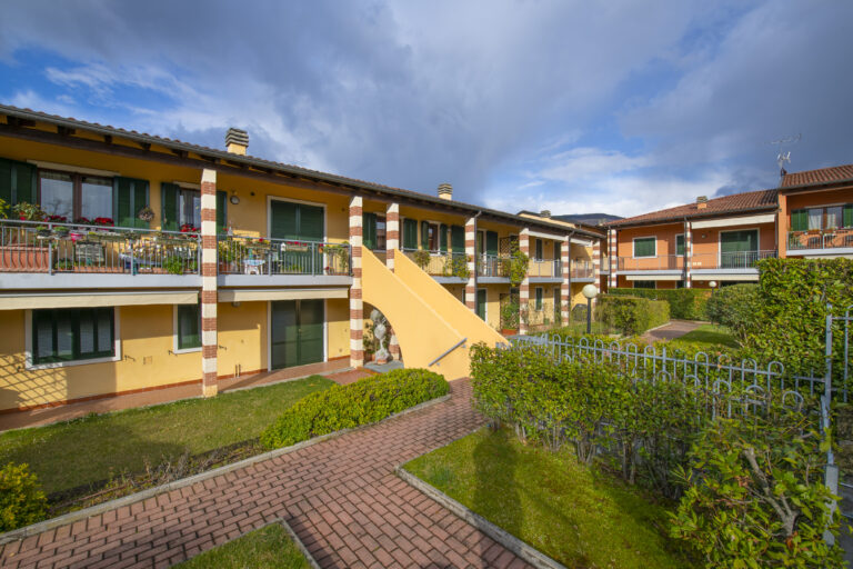Complejo residencial con edificios amarillos, balcones con plantas, caminos de ladrillo y setos verdes, cielo parcialmente nublado.
