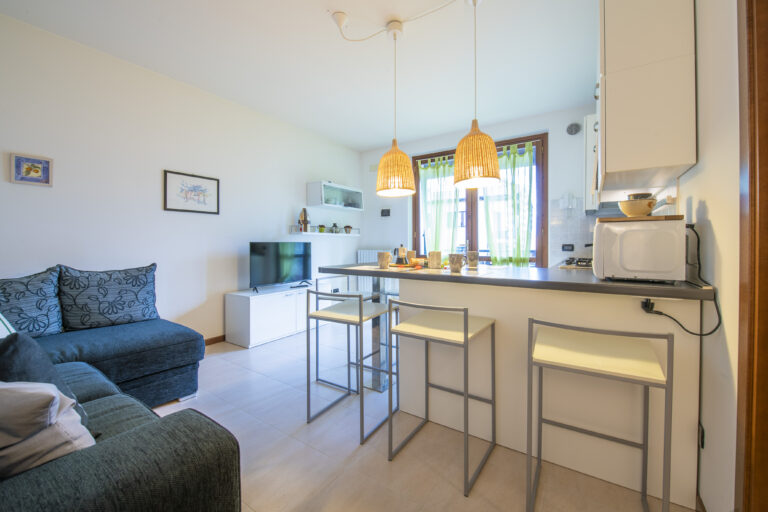 Sala de estar con sofá azul, barra de cocina con taburetes, lámparas colgantes, televisor y ventana con cortinas verdes.