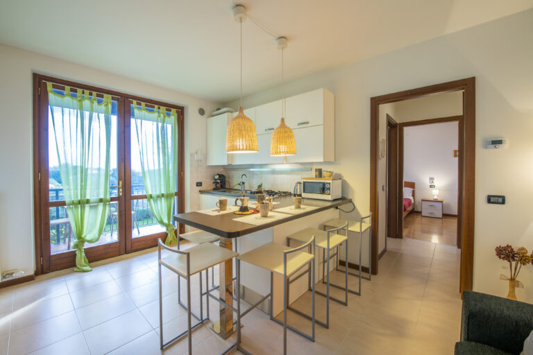 Cocina moderna con barra, sillas altas, luces colgantes, ventanales con cortinas verdes y vista al exterior. Habitación al fondo.