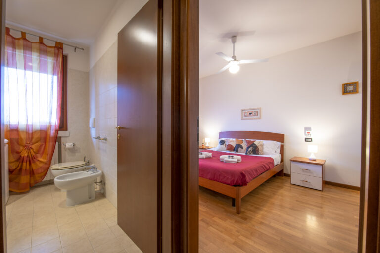 Vista de un dormitorio y baño. Cama con sábanas rojas, mesita de noche, ventilador, bidé y cortina naranja en el baño.