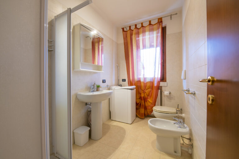 Baño con azulejos beige, ducha, lavabo con espejo, lavadora, ventana con cortinas naranjas, inodoro y bidé.