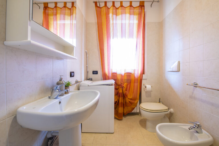 Baño pequeño con lavabo, inodoro y bidé blancos. Lavadora y cortinas naranjas traslúcidas en la ventana. Baldosas beige.
