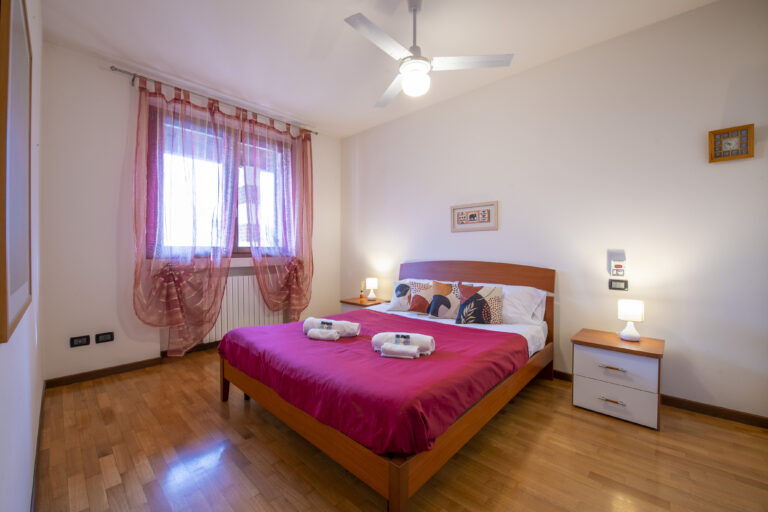 Habitación con cama matrimonial y edredón rojo, dos mesitas de noche con lámparas, cortinas rosadas y ventilador en el techo.