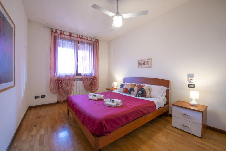 Habitación con cama de matrimonio cubierta por edredón rojo, lámparas de noche, cortinas rosas y ventilador en el techo.