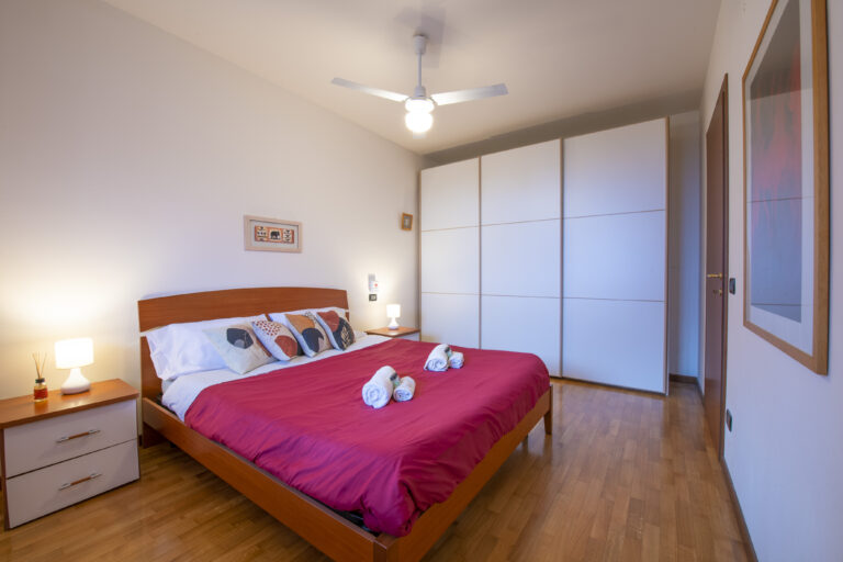 Habitación con cama de madera, edredón rojo, cojines decorativos, lámpara de mesa, armario blanco y suelo de parqué.