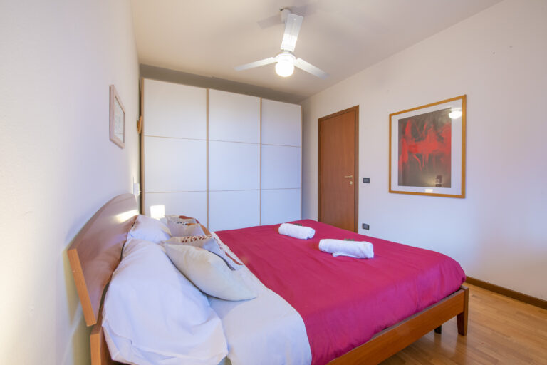 Habitación con cama de sábanas rosas, armario blanco, ventilador de techo y cuadro rojo en la pared. Decoración moderna.