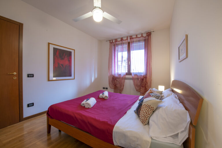 Habitación con cama de sábanas rojas, almohadas decorativas, luz de lámpara, ventana con cortinas rosas y cuadros en las paredes.