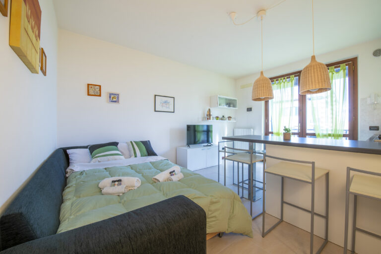 Sala moderna con cama, sofá oscuro, decoraciones en la pared, barra alta con taburetes, lámparas de mimbre y luz natural abundante.