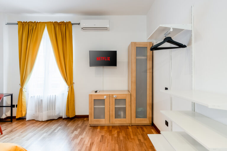 Sala con piso de madera, cortinas amarillas, televisor con Netflix, aire acondicionado, mueble de madera y estantería blanca.