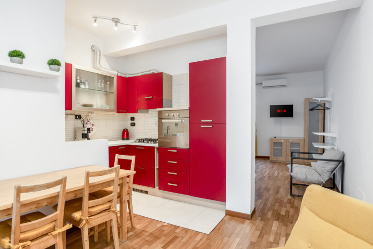 Cocina roja con mesa de madera y sillas. Sala adyacente con TV, sofá gris, aire acondicionado y suelo de parquet.