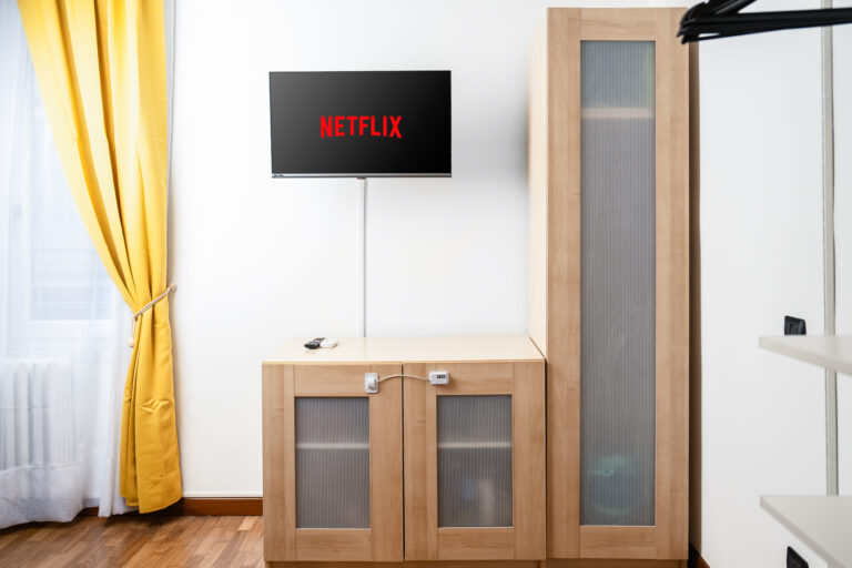 Sala con televisor mostrando el logo de Netflix, muebles de madera clara, cortinas amarillas y suelo de madera.