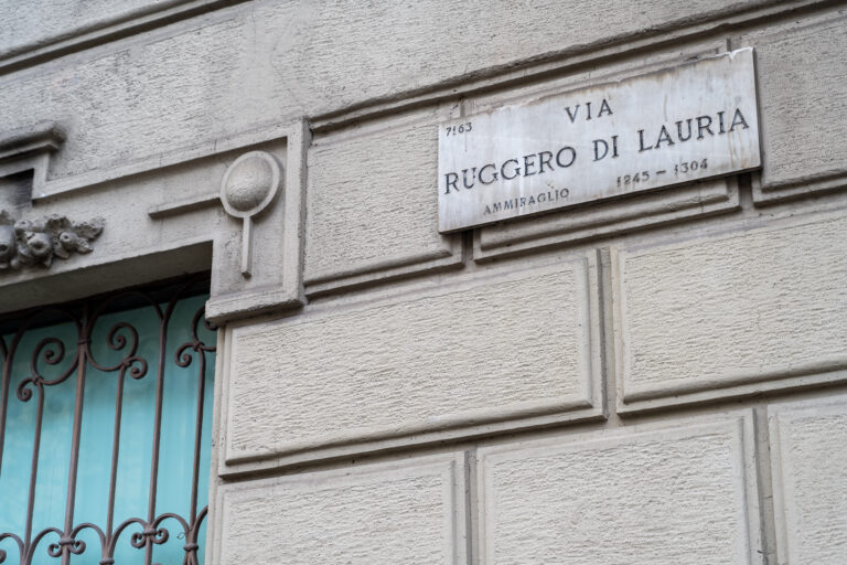 Pared de edificio con placa que dice "VIA RUGGERO DI LAURIA", ventana con rejas de hierro forjado y fondo azul claro.