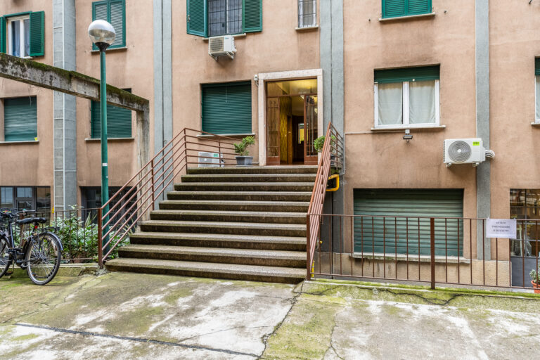 Entrada de edificio con escaleras de cemento, barandas de metal, plantas en macetas y puertas de vidrio. Ventanas verdes y paredes beige.