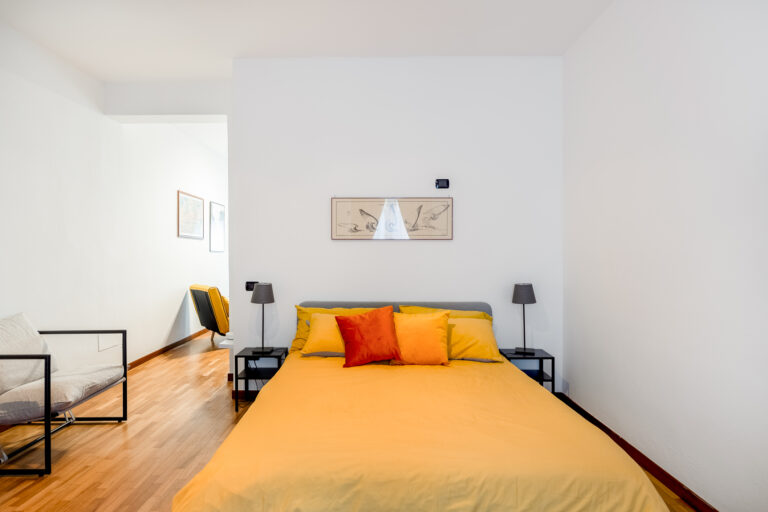 Dormitorio minimalista con cama de sábanas amarillas, cojines naranjas, lámparas negras y decoración de pared. Suelo de madera y pasillo al fondo.