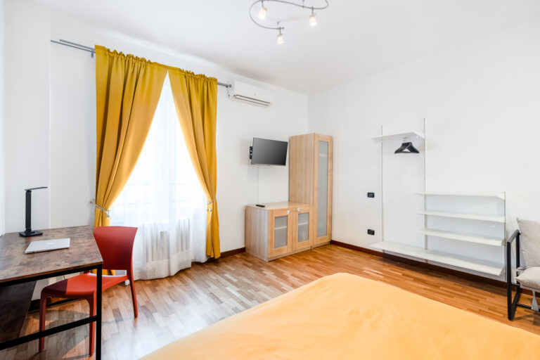 Habitación moderna con cortinas amarillas, cama con colcha naranja, escritorio con silla roja, mueble de madera, televisor y estantería.