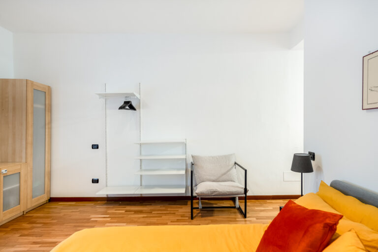 Habitación con suelo de madera, cama con ropa amarilla, silla gris, estantería blanca y lámpara de pie negra. Muebles modernos y minimalistas.