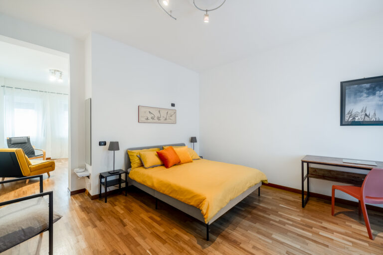 Habitación luminosa con cama de edredón amarillo, cojines de colores, escritorio de madera, sillas modernas, y suelos de madera.